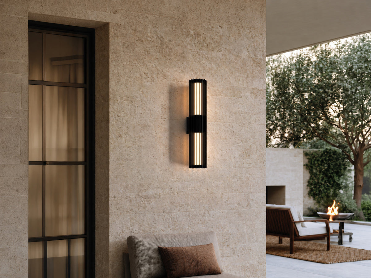 Applique extérieure cylindrique noire à lames, taille Moyen, allumée sur un mur en pierre claire au-dessus d’une terrasse avec cheminée extérieure