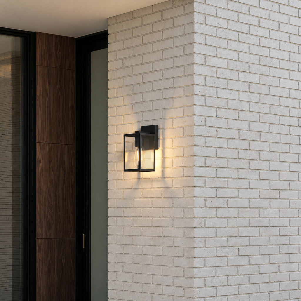 Applique lanterne noire allumée en blanc chaud sur un pilier en briques claires, à côté d’une porte d’entrée en bois