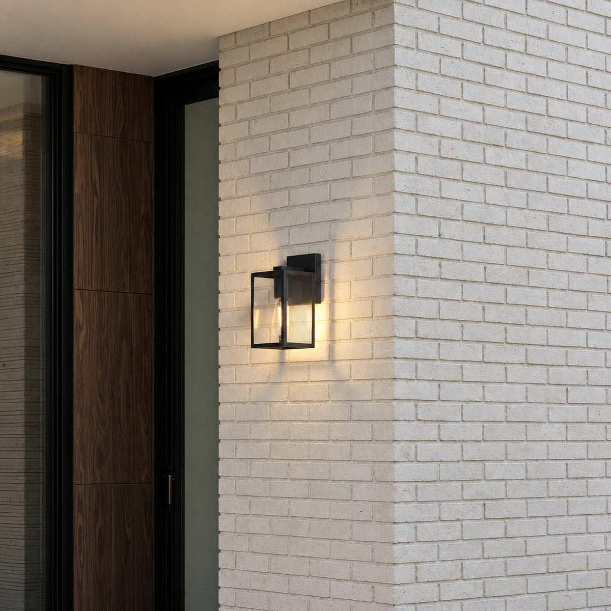 Applique lanterne noire allumée en blanc chaud sur un pilier en briques claires, à côté d’une porte d’entrée en bois