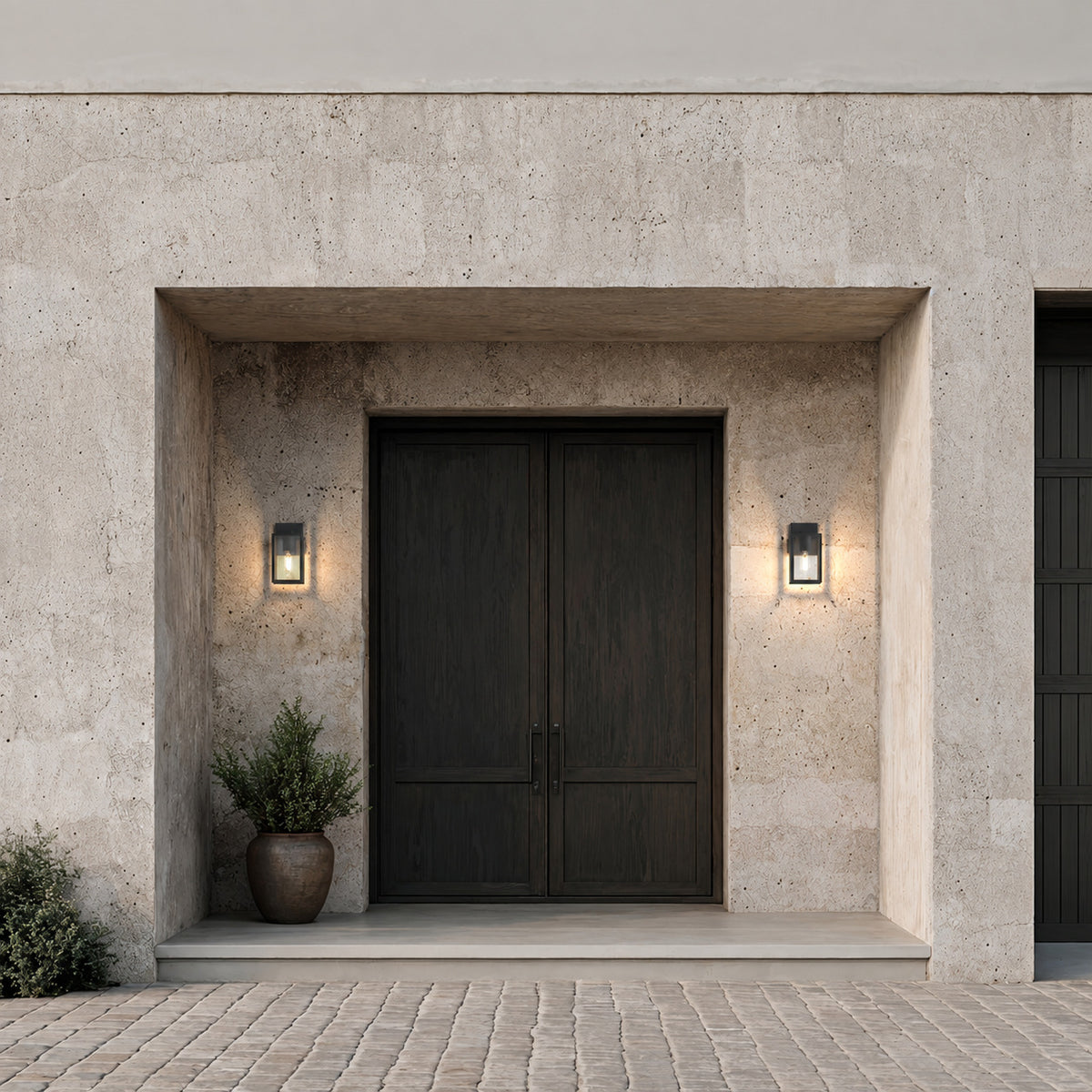 Deux appliques lanternes noires allumées de part et d’autre d’une double porte d’entrée en bois sombre, façade en pierre claire