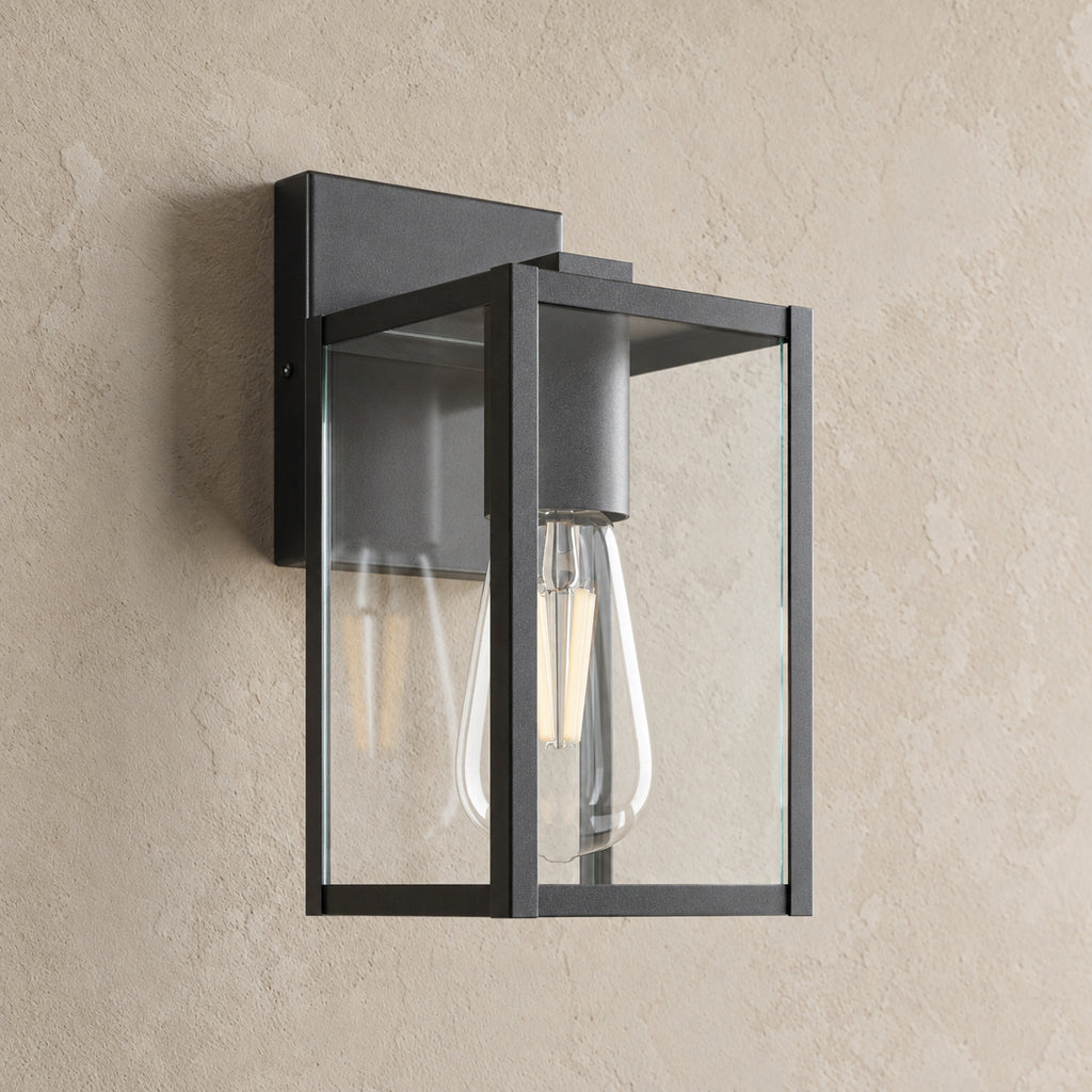 Applique extérieure lanterne noire, cage de verre transparent et ampoule LED de style filament, sur un mur enduit beige
