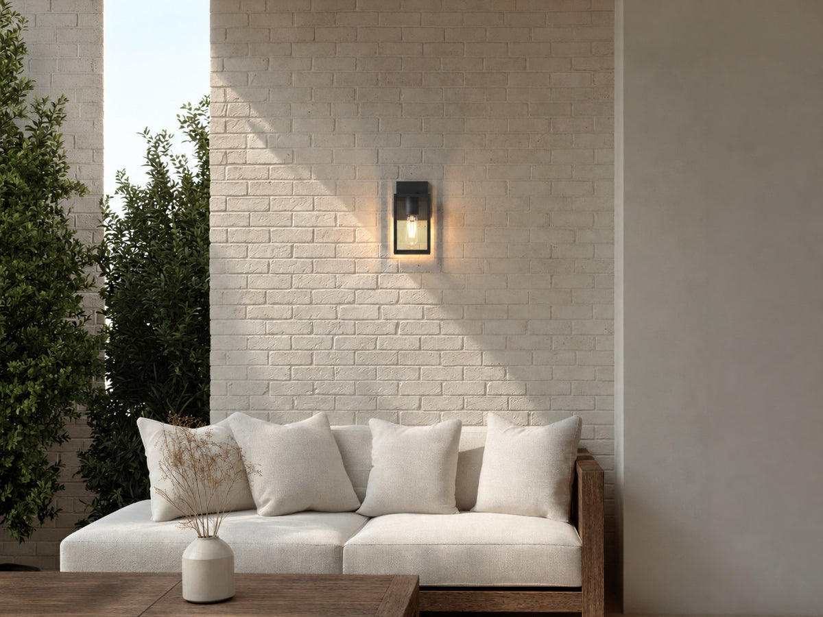 Applique lanterne noire allumée en blanc chaud sur un mur en briques peintes, au-dessus d’un canapé de terrasse
