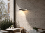 Applique murale horizontale noire allumée sur un mur de briques peintes, au-dessus d’une terrasse avec tables et chaises