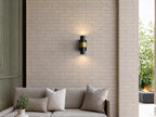 Applique extérieure noire et dorée allumée au-dessus d’un canapé de terrasse, sur un mur en brique claire
