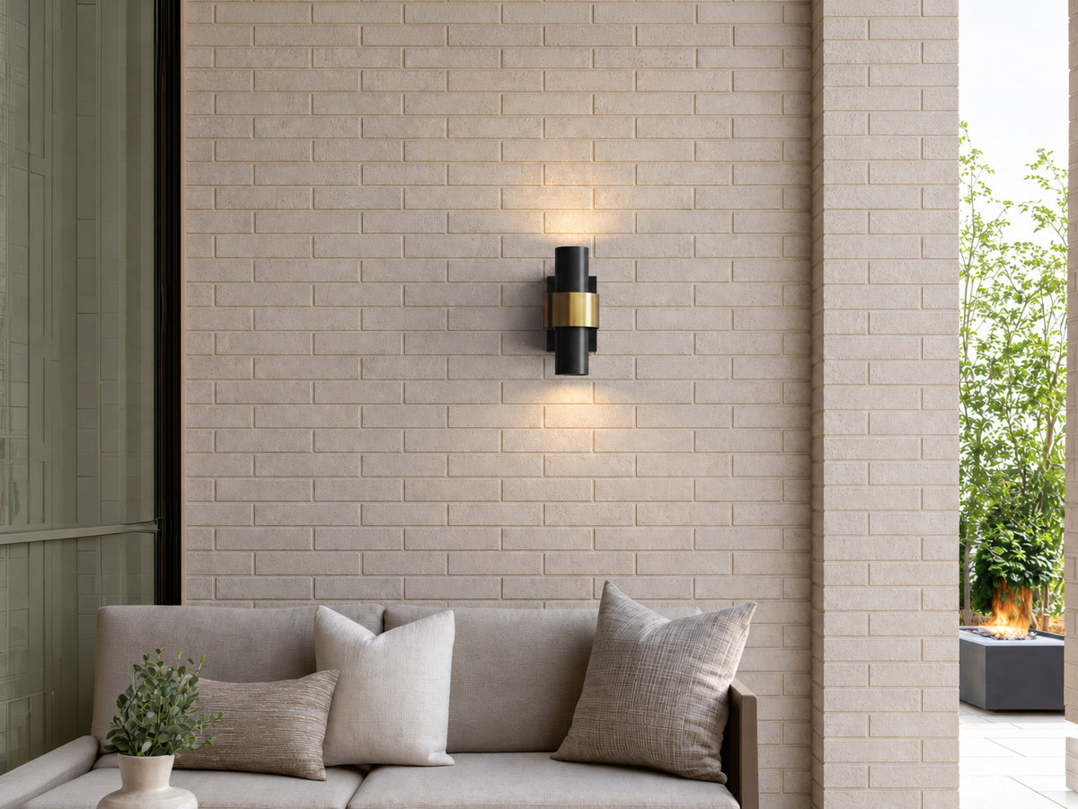 Applique extérieure noire et dorée allumée au-dessus d’un canapé de terrasse, sur un mur en brique claire