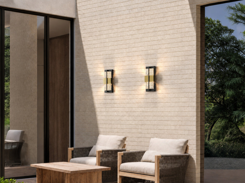 Deux appliques extérieures noires et dorées allumées sur un mur en briques claires, au-dessus d’une terrasse avec fauteuils