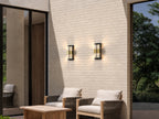Deux appliques extérieures noires et dorées allumées sur un mur en briques claires, au-dessus d’une terrasse avec fauteuils