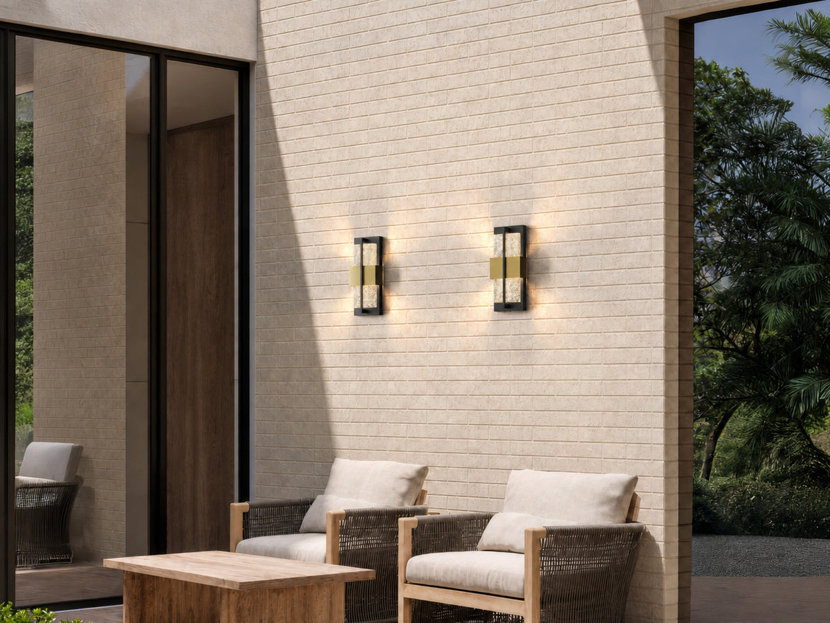 Deux appliques extérieures noires et dorées allumées sur un mur en briques claires, au-dessus d’une terrasse avec fauteuils