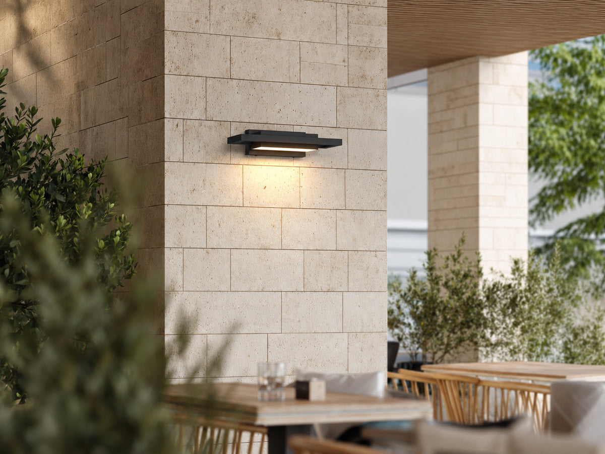 Applique extérieure noire à plans décalés allumée sur un pilier en pierre claire, au-dessus d’une terrasse avec tables de repas