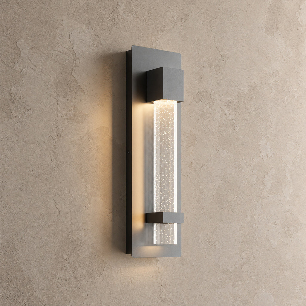 Applique murale extérieure noire allumée, colonne de verre bullé à lueur étoilée, vue de trois quarts sur un mur beige