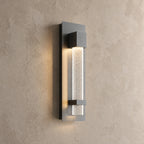 Applique murale extérieure noire allumée, colonne de verre bullé à lueur étoilée, vue de trois quarts sur un mur beige
