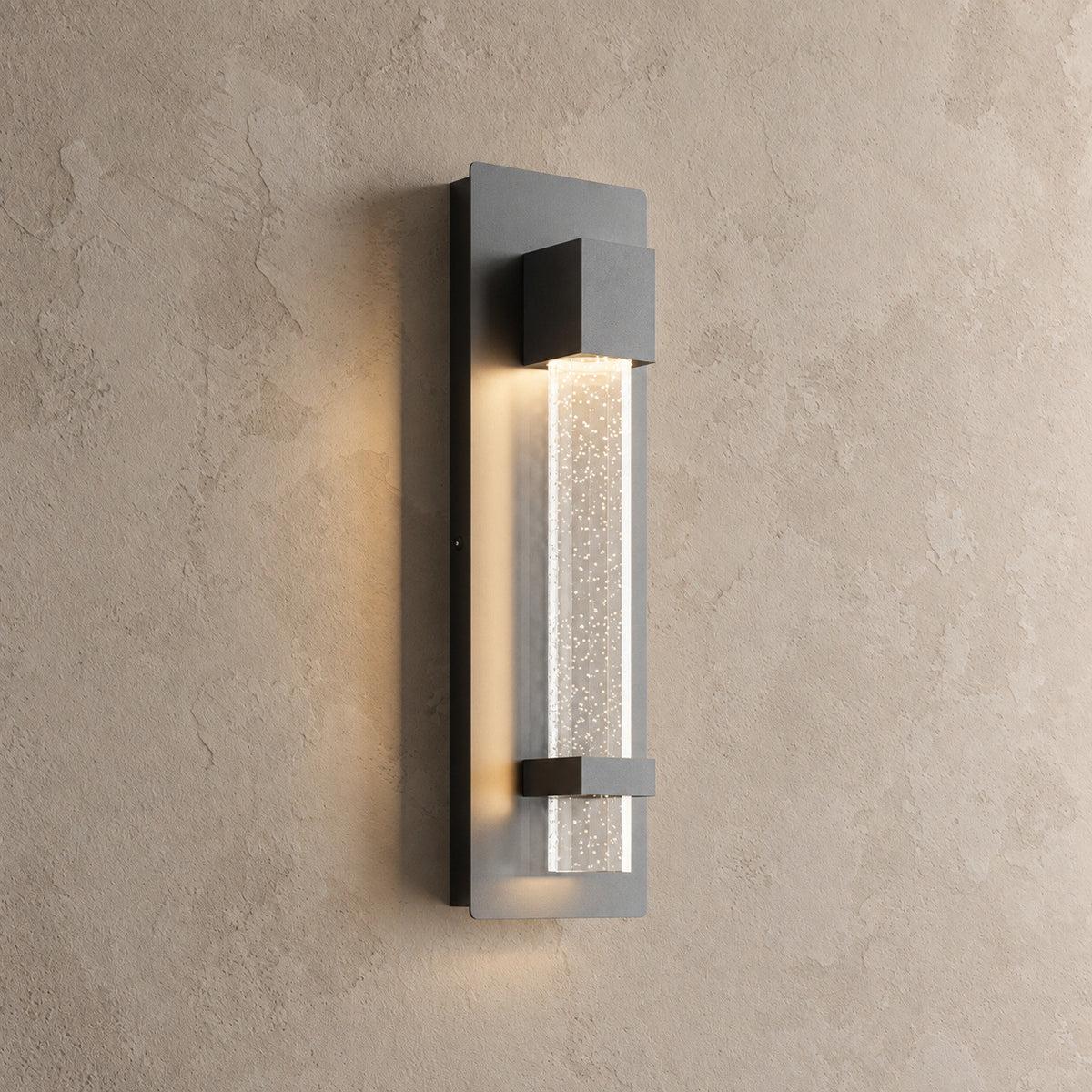 Applique murale extérieure noire allumée, colonne de verre bullé à lueur étoilée, vue de trois quarts sur un mur beige
