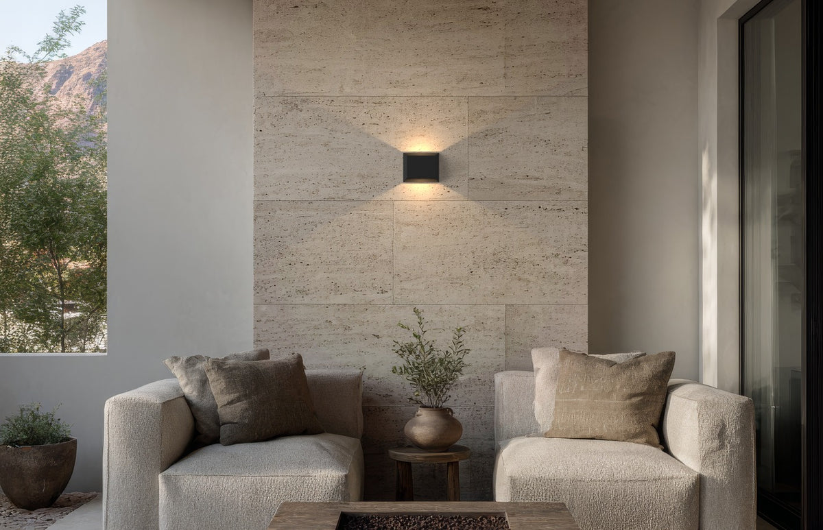 Applique extérieure carrée noire allumée sur un mur en pierre, au-dessus d’un salon de terrasse