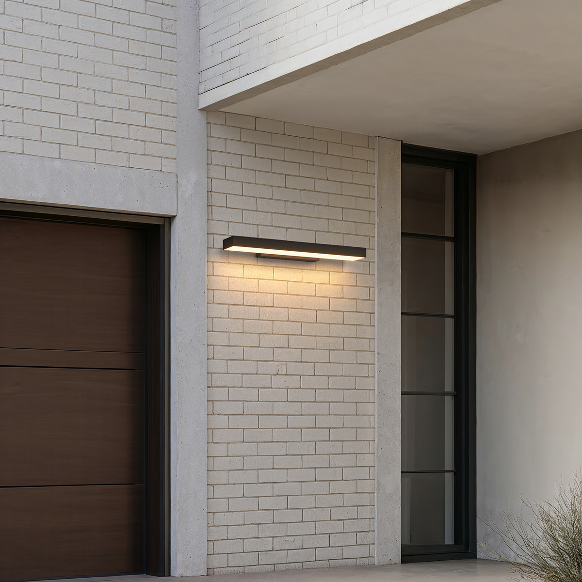 Applique extérieure horizontale noire 60 cm allumée sur un mur de briques blanches, entre une porte de garage et une entrée vitrée