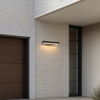 Applique extérieure horizontale noire 40 cm allumée sur un mur de briques blanches, entre une porte de garage et une entrée vitrée