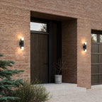 Deux appliques extérieures noires modèle double allumées en haut et en bas autour d’une porte d’entrée en bois, façade en brique