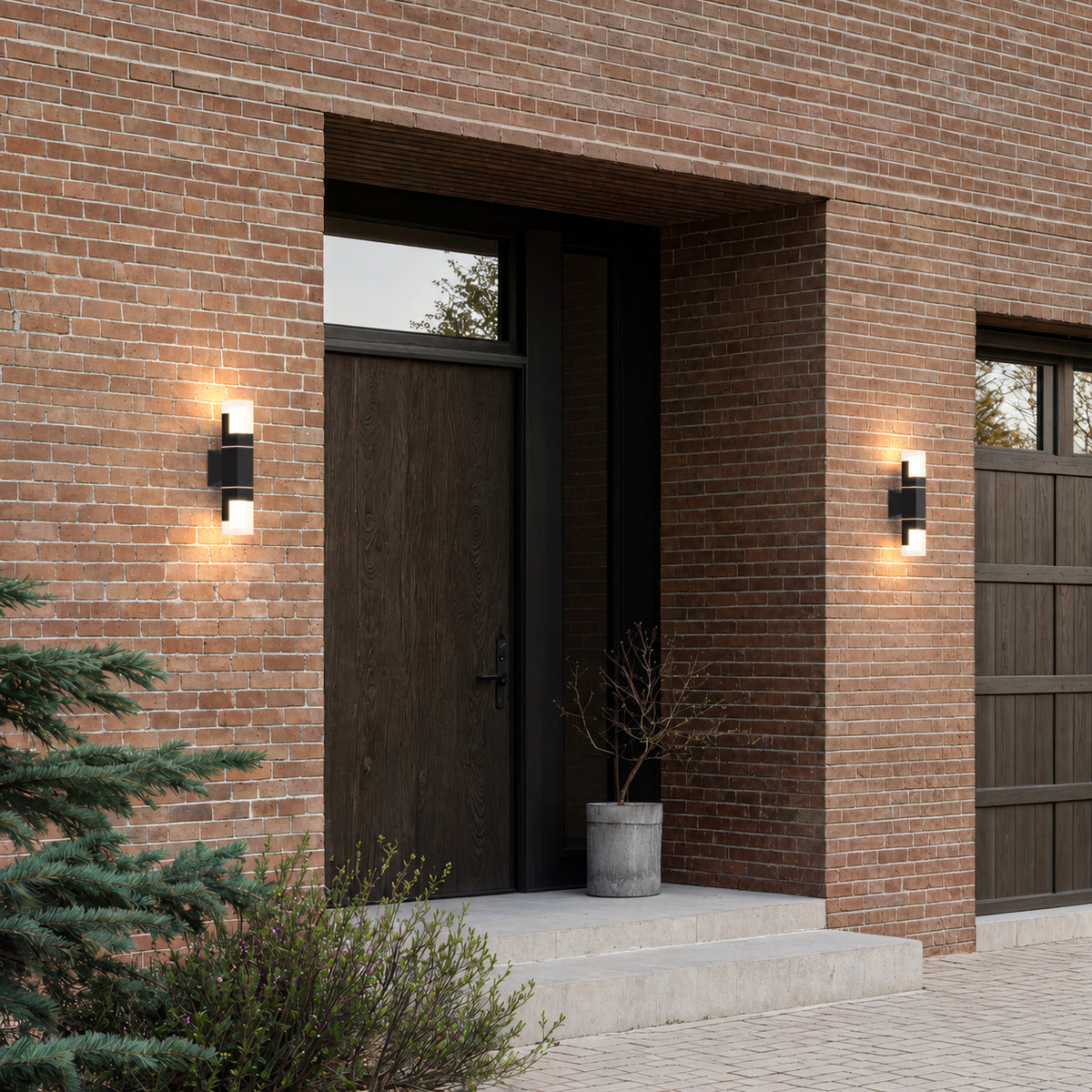 Deux appliques extérieures noires modèle double allumées en haut et en bas autour d’une porte d’entrée en bois, façade en brique