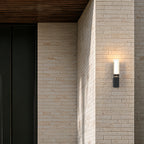 Applique extérieure noire modèle simple allumée sur une façade en brique claire, près d’une porte sombre