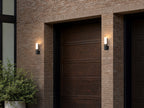 Deux appliques extérieures noires modèle simple allumées de part et d’autre d’une porte de garage en bois, façade en brique