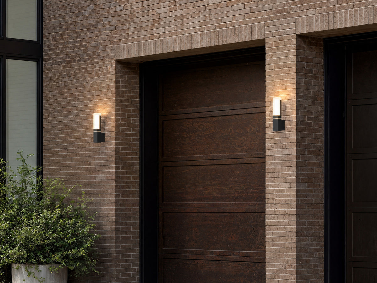 Deux appliques extérieures noires modèle simple allumées de part et d’autre d’une porte de garage en bois, façade en brique