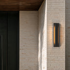 Applique extérieure LED lame verticale noire allumée sur une façade en brique claire, sous un auvent en bois