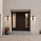 Paire d’appliques extérieures noires allumées de part et d’autre d’une porte d’entrée en bois sombre, façade en brique claire